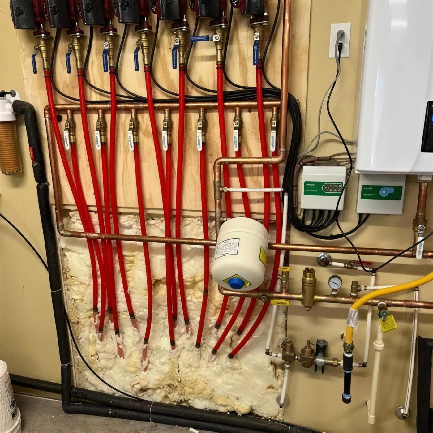 A PEX manifold with labelled shut-off valves feeding red PEX lines, next to a wall-mounted tankless water heater and an expansion tank.
