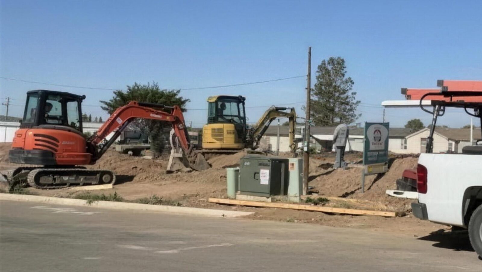 A new-construction lot in Alamogordo with two excavators, open trenching and a utility pedestal, under a clear blue sky.